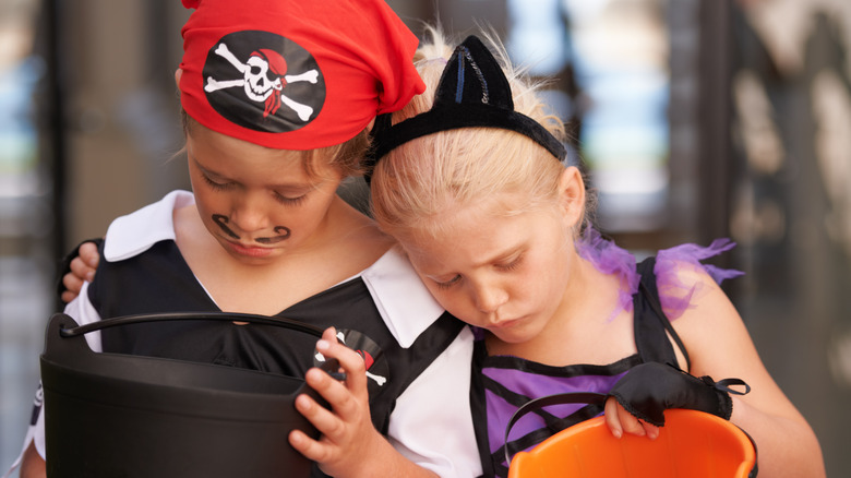Two kids in costumes look sadly into empty Halloween candy buckets.