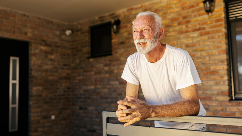 A happy retired man standing outside on the porch of a brick house.