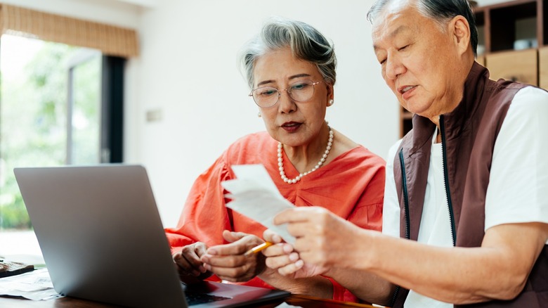 An older couple reviews receipts and financial paperwork beside a laptop at home