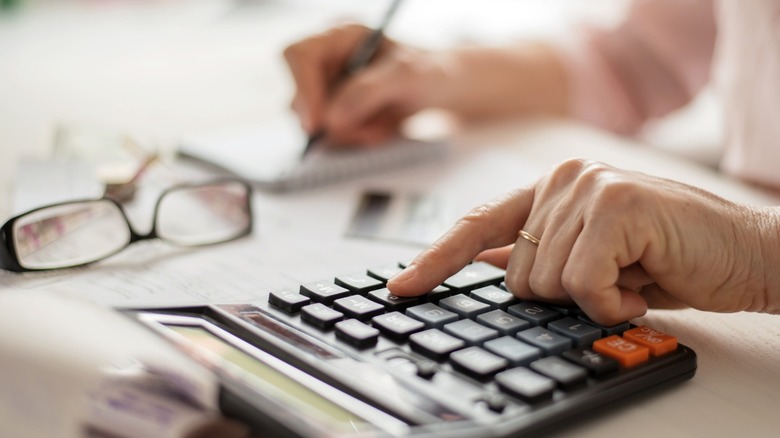 Close-up of a person using a calculator while another writes on paperwork in the background, with eyeglasses resting on the table