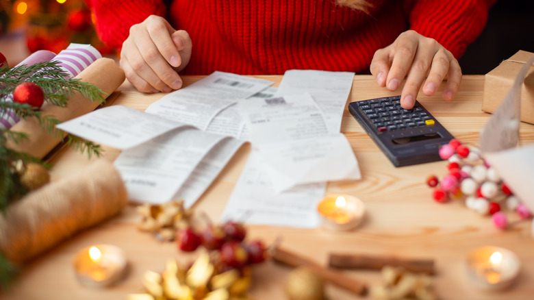 A woman sits at a table calculating her holiday spending with receipts and a calculator