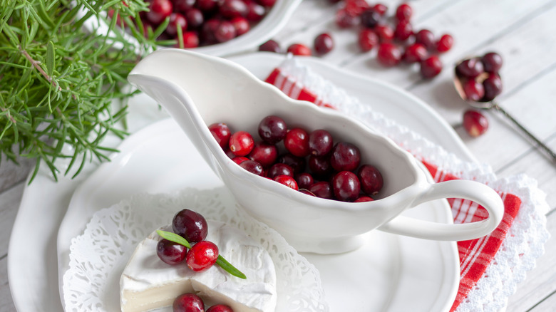 A gravy bowl full of fresh cranberries, with an out-of-focus plate of cranberries in background