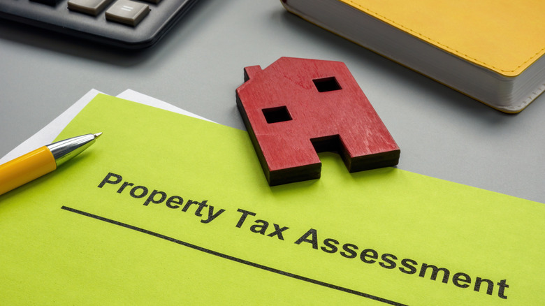 A green paper with "Property Tax Assessment" written on it in black with a small, red wood house and yellow pen on it with a calculator and yellow book in the background.