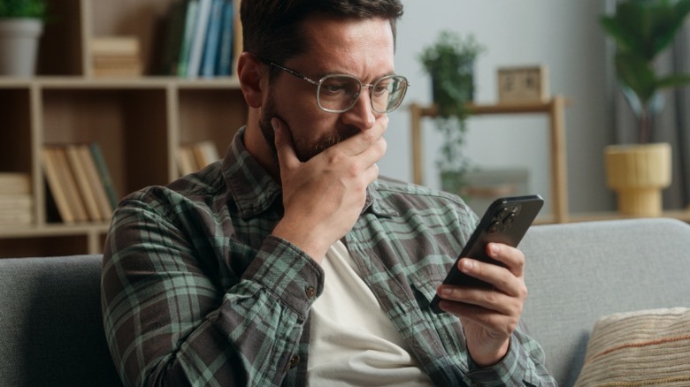 A man wearing glasses looking at his credit score on a phone with his hand over his mouth and an expression of concern.