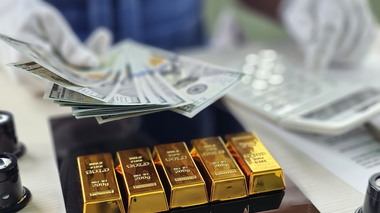 Person holding paper currency and using a calculator with gold bars sitting on table.