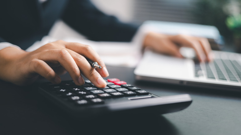 A close up of a hand using a calculator with the other hand using a laptop in the background.
