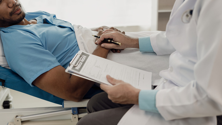 A young man lies on a bed in a hospital ward talking with a doctor