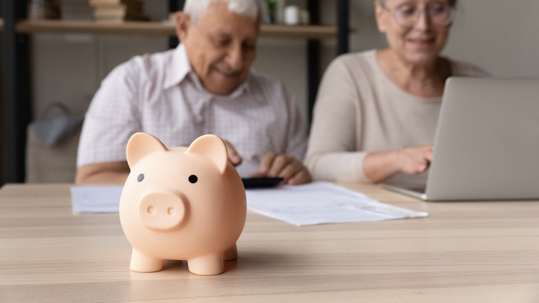 Focus on piggybank on the table, with an elderly couple make payment using e-bank on laptop in the background