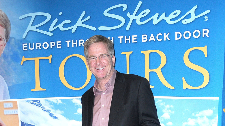 Rick Steves smiles in front of a promotional banner for one of his books