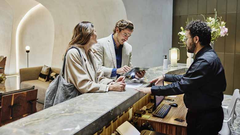 Medium shot of couple using digital tablet to check in at front desk in hotel lobby during business trip