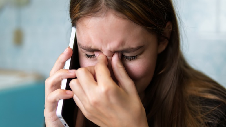 A distressed young woman holds a smartphone to her ear while pinching the bridge of her nose with her eyes closed
