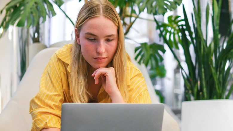 Young woman in a yellow shirt leaning on her hand while looking at a laptop indoors with plants in the background