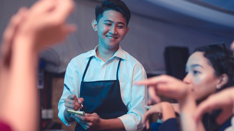 Smiling server in an apron taking an order from customers at a restaurant table.