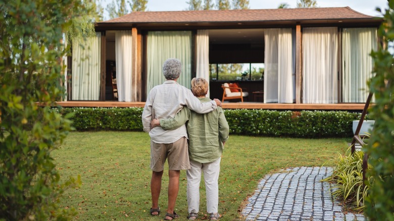 Older couple standing arm-in-arm on a lawn, looking toward a modern single-story home with large windows and a curved stone walkway