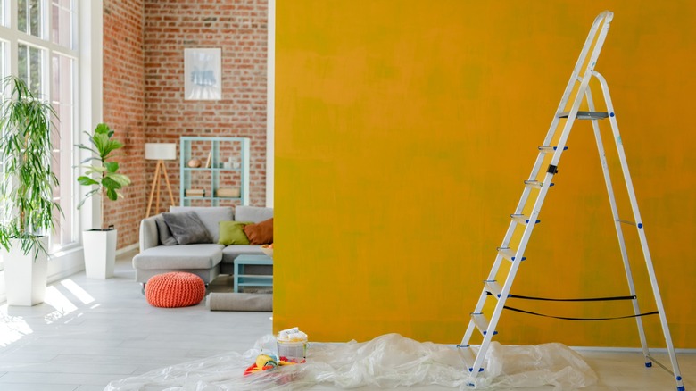 Bright living room mid-renovation with a yellow painted wall, ladder, paint supplies, and plastic sheeting on the floor