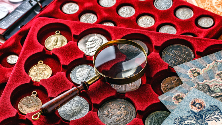 Collectible coins in a red velvet display with a magnifying glass on top and paper currency in the right corner.