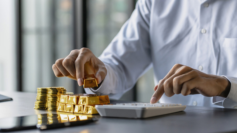 A person typing on a calculator while holding a gold bar