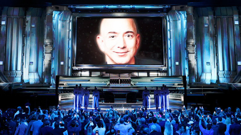 A headshot of Jeff Bezos floats in center stage at a conference attended by many people in a blue-toned room