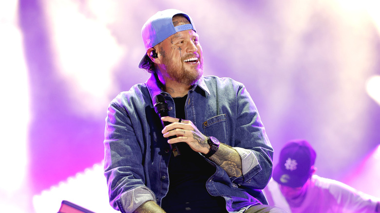 Jelly Roll smiles while performing with a microphone under purple and white stage lights