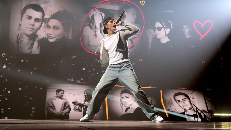 Justin Bieber performs onstage during his Justice World Tour, holding a microphone as photos and heart graphics appear behind him