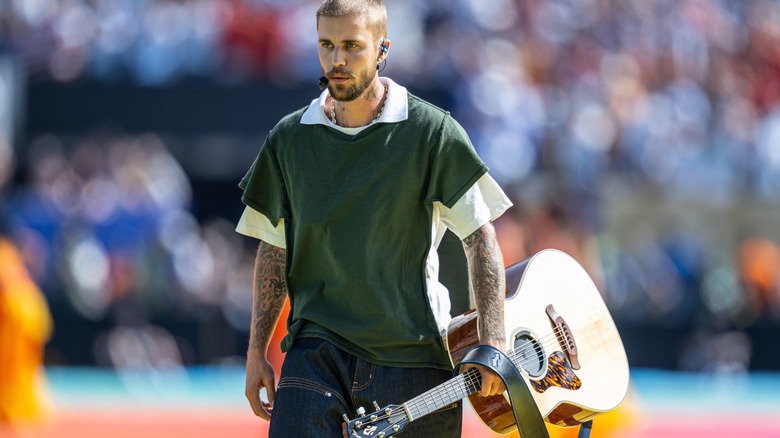 Justin Bieber carrying an acoustic guitar while performing onstage in front of a blurred stadium crowd