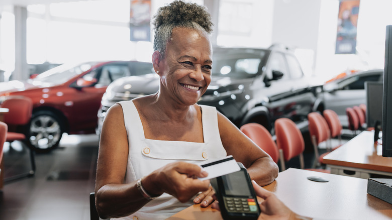 A smiling older woman uses a credit card to make a purchase at a car dealership