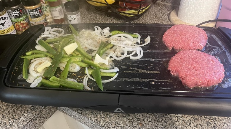 Onions and peppers grilling next to burger meat on an electric griddle.