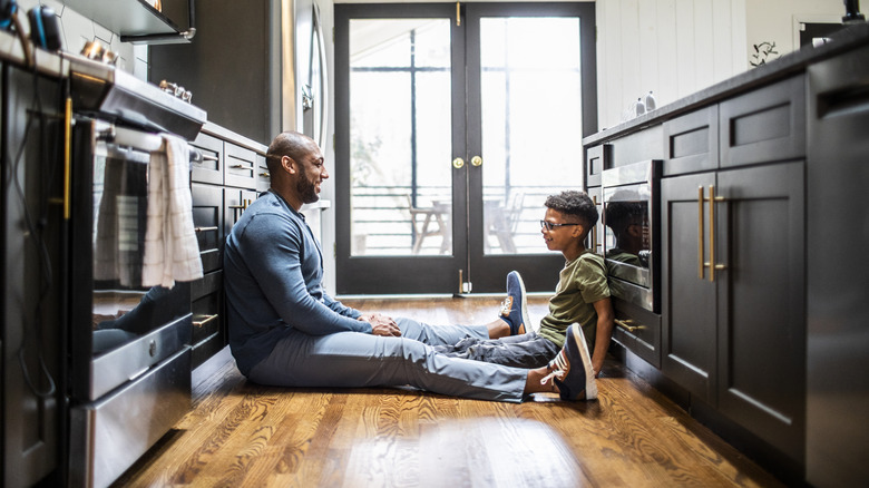 A man and a child sit on hardwood floor in a kitchen.