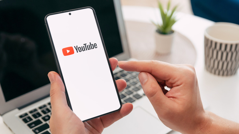 A closeup of a smartphone with the YouTube logo and a hand about to touch the screen with a laptop on a desk in the background.