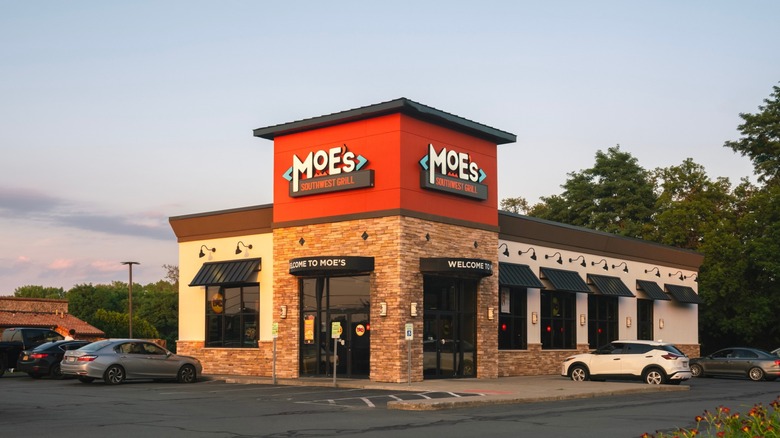 Exterior of a Moe's Southwest Grill restaurant with red signage, stone accents, black awnings, and cars parked outside