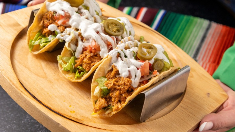 Three tacos filled with shredded meat, lettuce, tomatoes, jalapeños, and creamy white sauce, served on a wooden platter