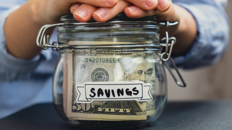Person with their hands on top of a glass savings jar with U.S. dollar banknotes inside