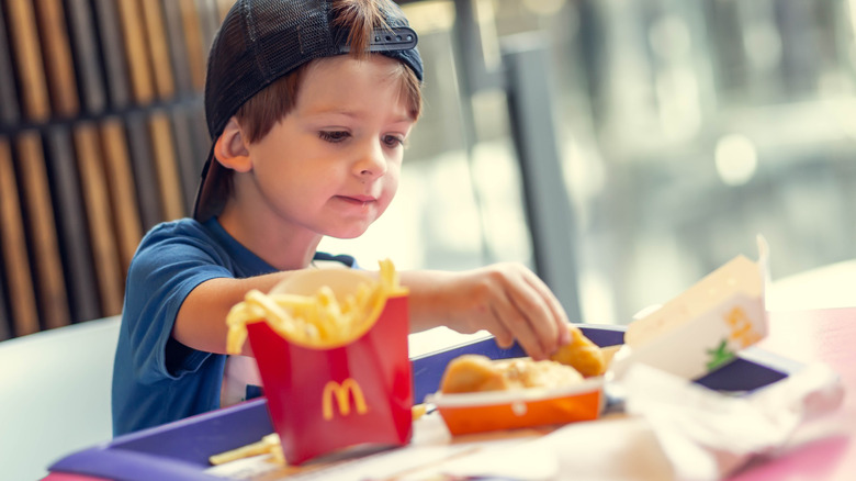 A young boy wearing a baseball hat backwards is eating McNuggets with fries at a McDonald's restaurant.