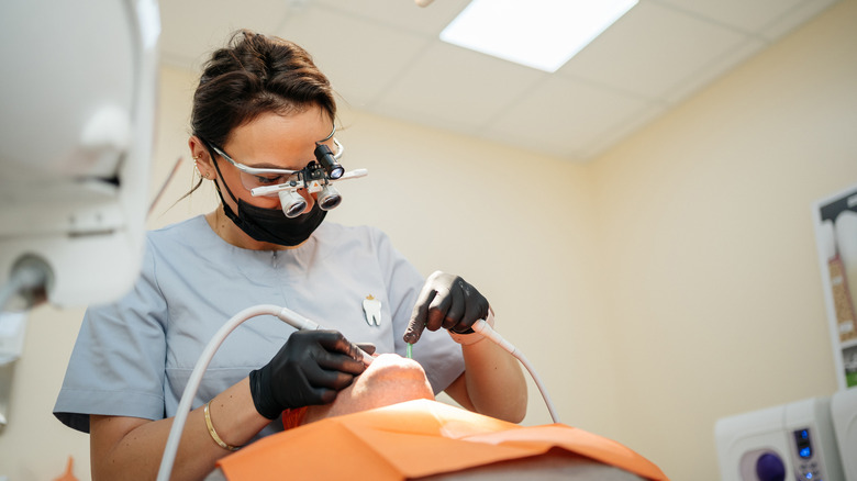 Patient lies in a chair while a dentist in binocular glasses treats teeth