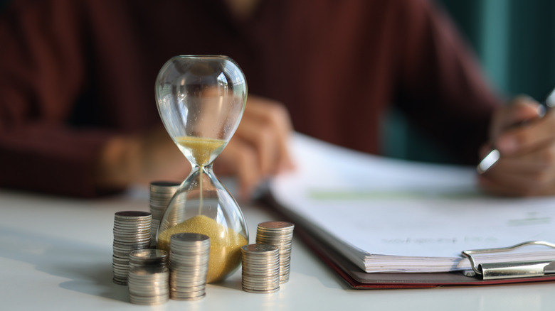 An hourglass next to coins on a desk, with a person reading documents in the background.