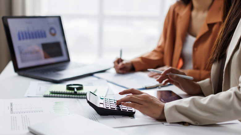 Two people sitting at a desk and using a calculator and a laptop to perform calculations