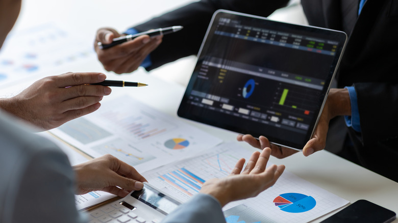 Hands holding tablet with financial data over desk covered in paperwork.