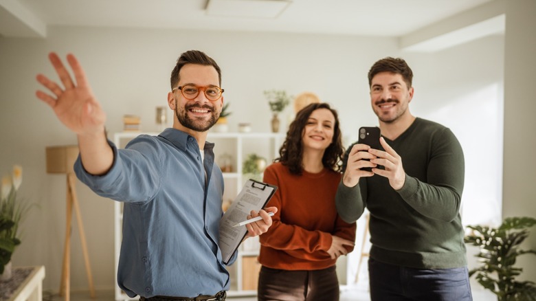 A smiling real estate agent gestures around a bright living room while a couple looks on, with one person holding a smartphone