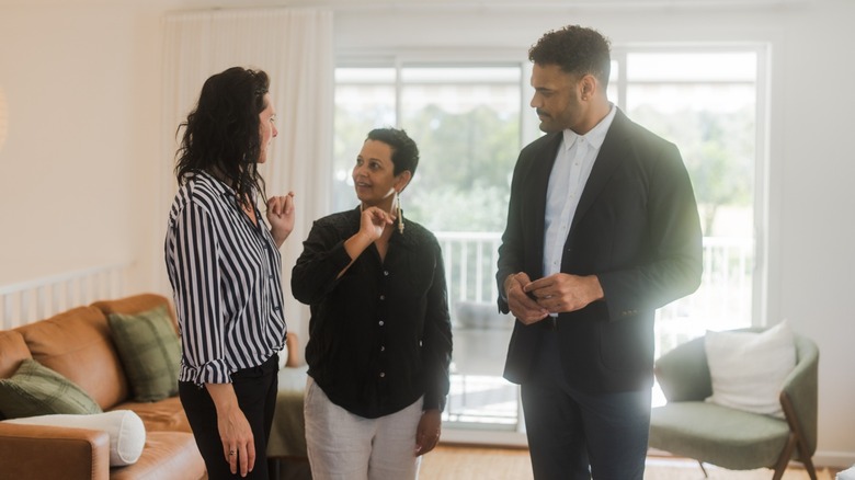 Three people talk while standing in a bright, furnished living room during a home viewing
