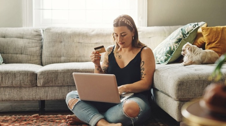 A young woman with tattoos sitting on a living room floor on a laptop while holding up a credit card.