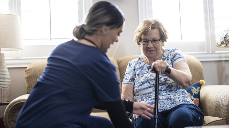 An elderly woman with a cane getting a checkup from a younger home health aide in her living room.