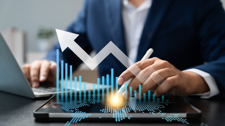 A close up of a hand holding a stylus using a tablet with an overlay of a 3D graph and an arrow pointing up with  the other hand in the background using a laptop.