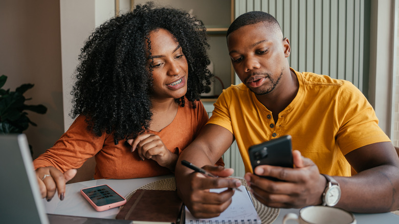 A young couple sitting at a table looking at a phone the man is holding with a laptop, phone, and pad of paper on the table in front of them.