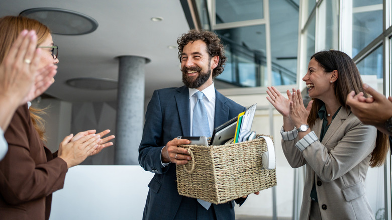Smiling businessman holding box with office supplies while surrounded by clapping peers