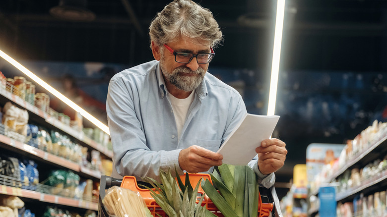 Smiling man reads over grocery list while leaning over shopping cart in store