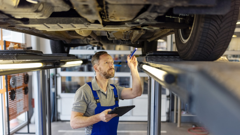Bearded mechanic with blue overalls and a grey collar shirt holds a thin blue flashlight as he checks the underside of a vehicle on a car lift.