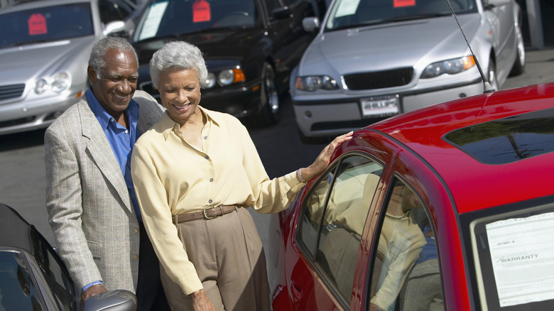 A smiling retired couple at a car lot looking at a red car with other vehicles in the background.