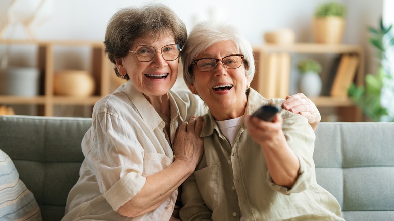 Two older female friends sitting on couch and pointing remote control