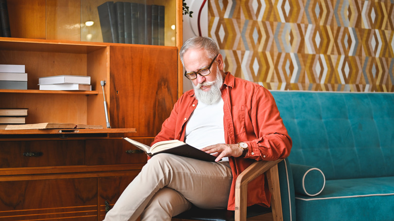 A relaxed mature man reading a book while seated in a chair in his home.