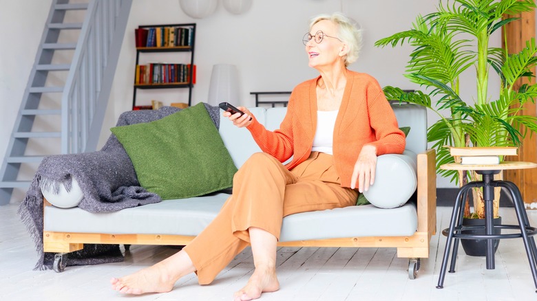 A smiling mature woman holding a tv remote while sitting barefoot on a couch.